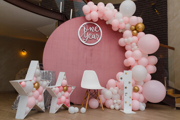 Beautiful festive decorations, pink and golden balloons arch, wooden stars, white chair and number one on wooden round background. Little 1 year old girl birthday party photo zone.