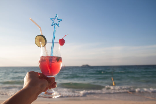 Woman's hand holding red color glass of summer drink with the beach on background