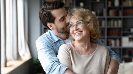 Loving young handsome man embracing from back and kissing happy older retired mother, showing care and devotion at home. Head shot affectionate grown son feeling appreciation to mature elderly mother.