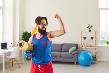 Funny athlete holding gym mat and dumbbells and showing his weak biceps after sport workout at home