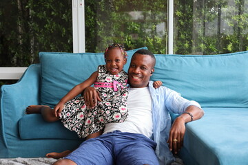 African American father playing with his little daughter who sitting on the couch while spending happy time together at home on sofa