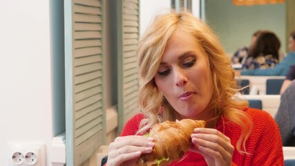 beautiful young woman in a red sweater eating a croissant in a cafe. blonde in a stylish cafe has a sandwich - Powered by Adobe