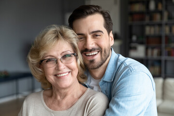 Fototapeta premium Head shot close up portrait of loving young bearded man cuddling smiling beautiful middle aged mature mother on eyeglasses. Loving different generations family looking at camera, enjoying weekend.