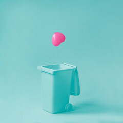 Pink heart falling in garbage bean against pastel blue background. Broken, fake, lost love. Minimal relationship concept.