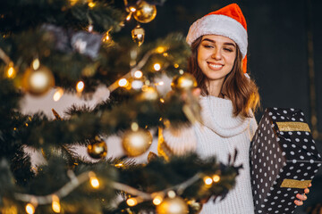 Pretty woman in warm sweater, socks and christmas hat, decorating new year tree and holding gif box