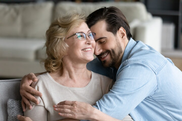 Obraz premium Young caring son cuddling happy elderly senior retired mother in eyeglasses, feeling thankful at home. Smiling 60s mature woman enjoying sincere trustful relations moment with grown up child at home.