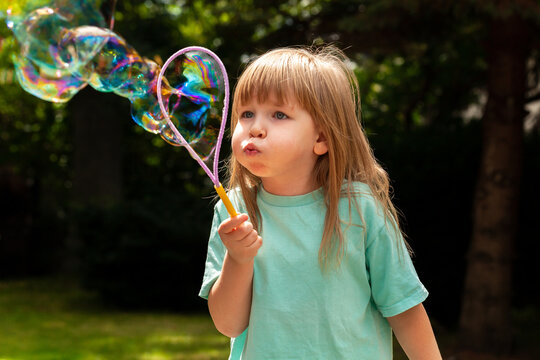 Little child, girl blowing huge bubbles alone, portrait outdoors. Young kid with puffed cheeks blows big bubbles outside, closeup shot Playing, fun activities for children, happiness concept lifestyle