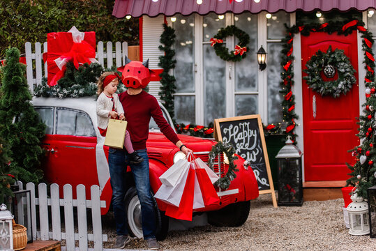 A young man in a red bull mask is holding a little baby, white packages. New Year's shopping, sale. The family stands by the car, trees in the snow decorated for Christmas. Girl kiss an ox. Copy space