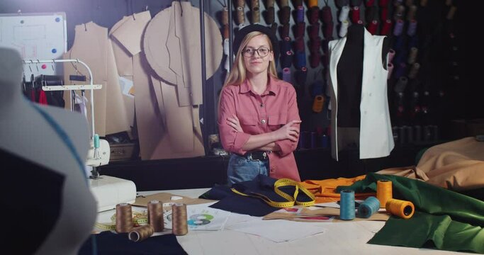Young Caucasian female clothes designer in hat standing in workshop at table. Cheerful beautiful woman tailor in glasses at her office in atelier, looking at camera. Working process. Job concept