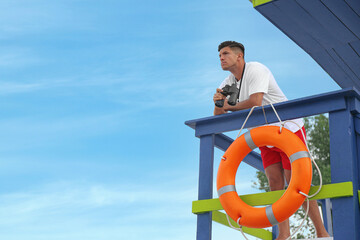 Male lifeguard with binocular on watch tower against blue sky - obrazy, fototapety, plakaty
