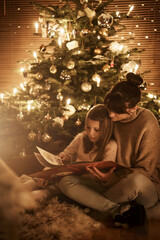 Child sits with mother in front of the Christmas tree and read a book together and look forward to...