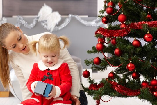 Little girl and mother sitting by christmas tree, opening presents.