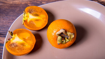 Persimmon cut in half and whole persimmon on a plate on a wooden table, close-up, copy space