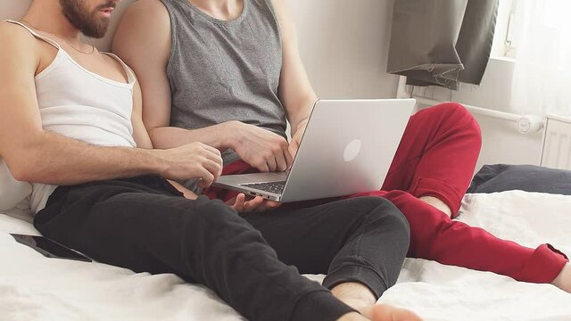European Gay lgbtq men couple using computer laptop at home. Young lovers watching movie lying on bed in bedroom at house.