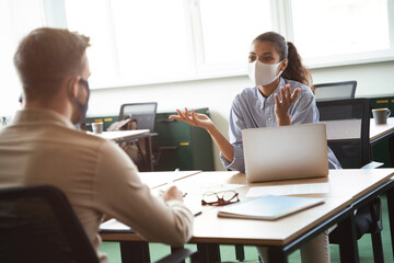 Two young diverse colleagues wearing protective face mask discussing something while working together in office during covid19 outbreak