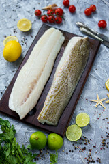 fillet of white fish two pieces on a wooden board on a gray background with knives lemons and tomatoes
