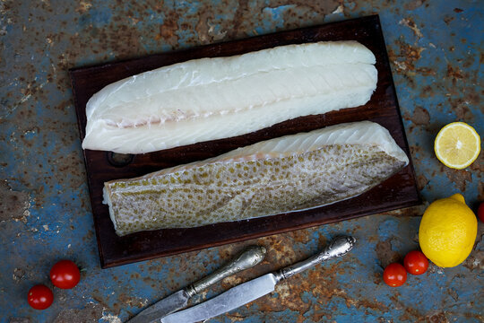 Fillet Of White Fish Two Pieces On A Wooden Board On A Blue Rusted Sheet Of Metal With Knives Lemons And Tomatoes
