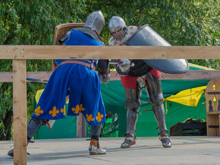 Two foot warriors in heavy medieval armor fight in the arena. Armed with swords. They are protected by iron helmets and shields. Historical reconstruction of medieval European knightly tournaments