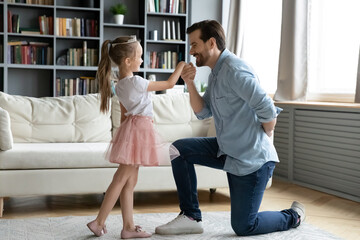 Full length gentle loving caring young father standing on knee, kissing hand of little princess dressed adorable child daughter, inviting for dance in living room, domestic hobby playtime activity.