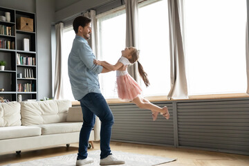 Full length energetic young father spinning laughing joyful little preschool child daughter, having fun together in modern living room. Smiling daddy lifting in air funny small kid girl at home.
