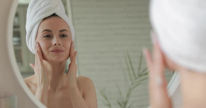 Happy lady in white towel standing in bathroom and applying cream on face. Young woman with bare shoulders using korean cosmetics for skin care.