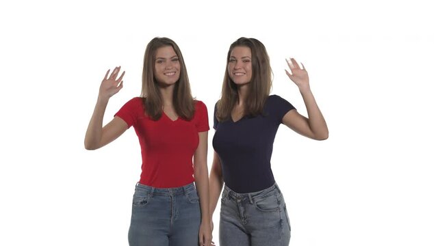 Portrait of smiling Caucasian twin sisters looking at camera and waving hands in greeting. Hello or goodbye gesture. Isolated on white background