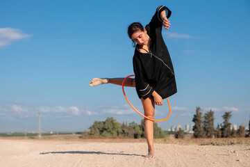 Female flow artist dancing with a Hula hoop outdoors, with one leg on the ground. Wide shot in natural morning sunlight. 