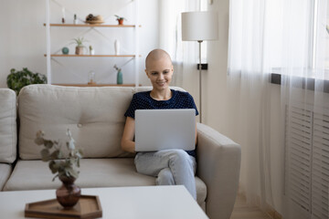 Smiling hairless woman using laptop, sitting on cozy couch in living room, happy young female enjoying leisure time with gadget, having fun online, chatting or shopping, watching video or movie