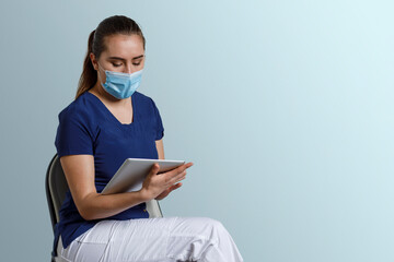 Mujer latina, con uniforme de enfermera usando cubrebocas y careta, sosteniendo una tableta digital en las manos, y un fondo color azul con espacio para texto