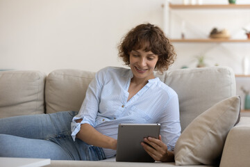 Close up smiling young woman lying on couch, using computer tablet, relaxing with gadget at home, happy female holding device, surfing internet, chatting or shopping online, enjoying leisure time