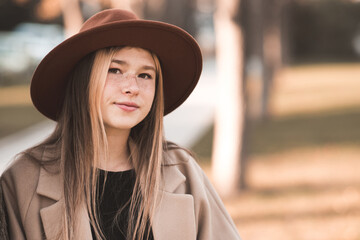 Beautiful blonde teen girl 13-14 year old wearing hat and jacket in park outdoors. Looking at camera. Teenagerhood. Fall season.