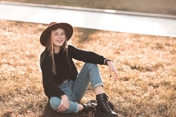 Smiling beautiful teen 13-14 year old wearing stylish clothes and hat sittin in park on green grass outdoors close up. Looking at camera. Childhood. Teenagerhood.