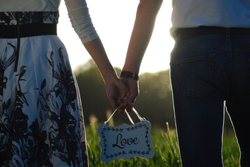 zakochani kobieta i mężczyzna spacerując po łące w wysokiej trawie podczas zachodu słońca,  wspólnie trzymając, niosą  w dłoniach białą tabliczkę z napisem love