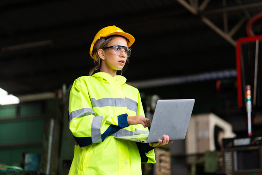 Female Quality Control Inspector Checking Workers At Factory. Woman Engineer With Yellow Hard Hat Helmet Working On Laptop Computer Inside Industrial Manufacturing Factory