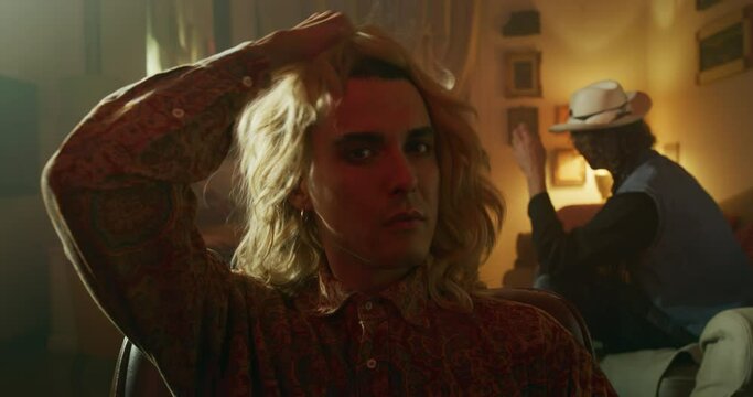 Cinematic shot of young homosexual male gay is taking off wig and smiling in camera on a background of his friends are having fun with camera. Concept of gender expression, identity and diversity.