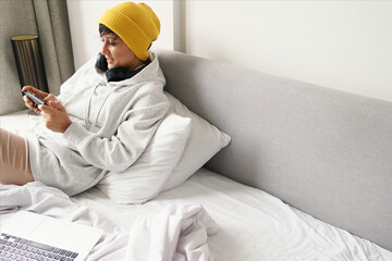 Young woman in yellow hat, headphones is watching screen of smartphone and laptop on bed. Technology