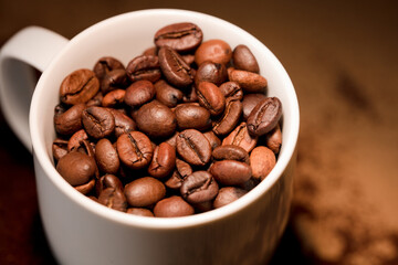 Coffee beans, with ground (selective focus on coffee beans) and white ceramic cup, brown background.