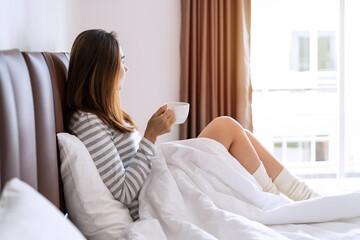 Young woman relaxing at cozy home atmosphere and look out the window on the bed with cup of coffee in the morning