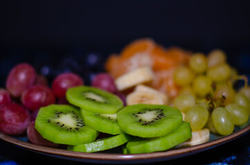 Fruits, grapes, tangerines, bananas, kiwi in a blue porcelain plate. black wooden floor, fruit plate