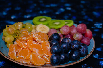 Fruits, grapes, tangerines, bananas, kiwi in a blue porcelain plate. black wooden floor, fruit plate