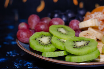 Fruits, grapes, tangerines, bananas, kiwi in a blue porcelain plate. black wooden floor, fruit plate