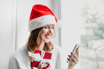 A woman in a New Year's hat holds a red gift box smiles and makes a video call while sitting on the windowsill in her room. Coronivirus quarantine, distance video calls