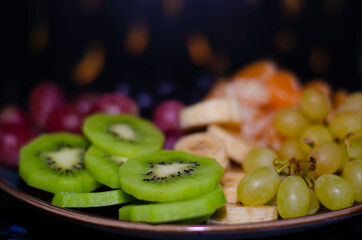 Fruits, grapes, tangerines, bananas, kiwi in a blue porcelain plate. black wooden floor, fruit plate