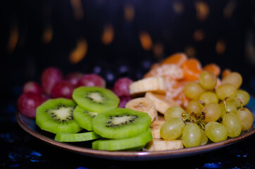 Fruits, grapes, tangerines, bananas, kiwi in a blue porcelain plate. black wooden floor, fruit plate