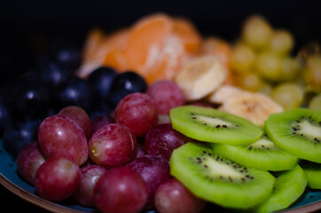Fruits, grapes, tangerines, bananas, kiwi in a blue porcelain plate. black wooden floor, fruit plate