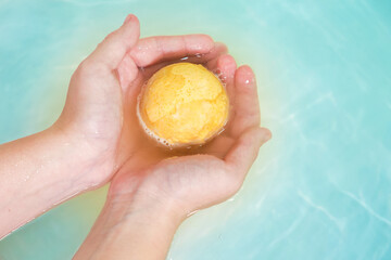 Salt bath bomb. Orange bubbling ball of bath salt in the hands on blue water background. Bomb for...