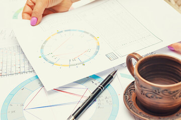 Workplace of a modern astrologer. Printouts of astrological charts, charts, tables, diagrams lying on the table. Hands of a woman astrologer. A cup on a saucer.