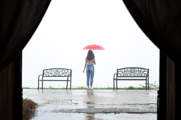 Young Asian Woman holding red with white dot umbrella standing between chair and contemplates under rain with overcast sky