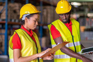 Woman worker holds a clipboard with a man holding tablet and checks goods in the automotive parts warehouse distribution center. Both people wear a safety helmet and vest. In background shelves