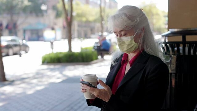 Wearing a mask in compliance with COVID19 mandates a mature woman sitting in an outdoor urban environment looks at her smartphone and drinks coffee.